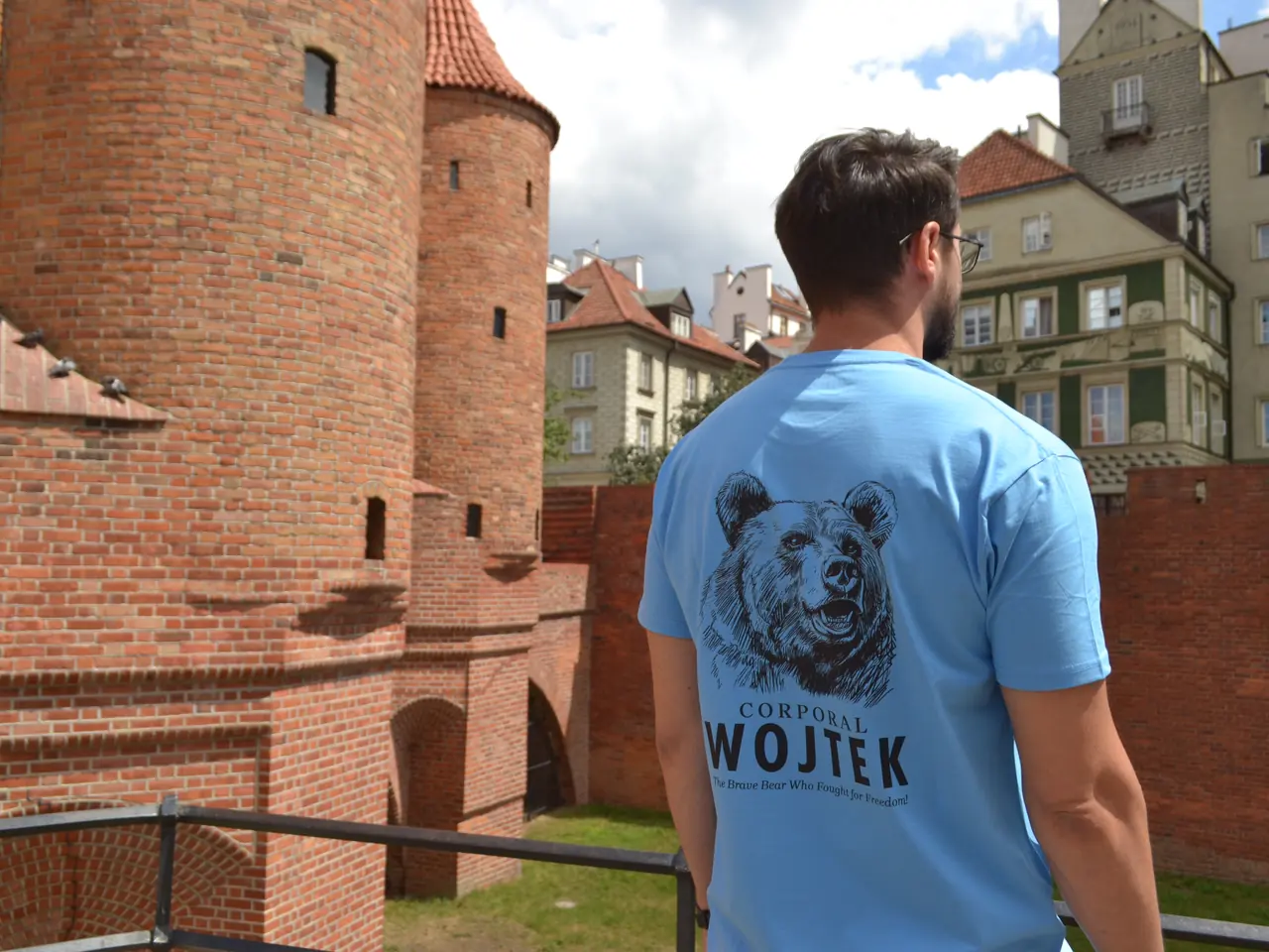 Mężczyzna w błękitnej koszulce Corporal Wojtek stoi tyłem przy Barbakanie w Warszawie. Na plecach duży nadruk z niedźwiedziem i napisem Corporal Wojtek.